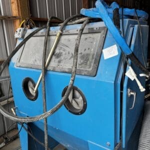 Blue industrial sandblasting cabinet with a viewing window, two glove ports, and tangled hoses in a cluttered workshop surface area