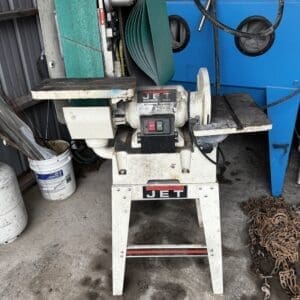 Jet brand benchtop belt sander on a white stand in a dusty workshop, with a green abrasive belt around the drum.