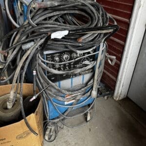 Blue welding machine tangled with thick black extension cords on a concrete floor in a workshop/garage setting