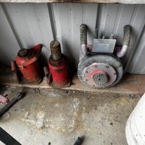 Old dusty workshop corner with red hydraulic jacks and a large round vintage tool leaning against a corrugated metal wall.