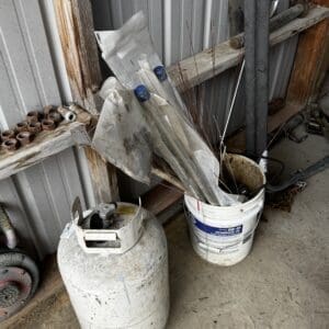 Cluttered shed corner with a large propane tank and a white bucket filled with thin metal rods and tools nearby.