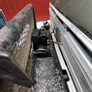 Close-up of a trailer bed with a winch and coiled cable on a diamond-plate surface, a rusted ramp leaning nearby, and a red wall in the background.
