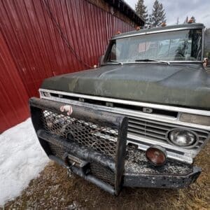Vintage Ford pickup with a rusted front bumper and grille guard parked on muddy snow beside a red corrugated wall barn exterior.