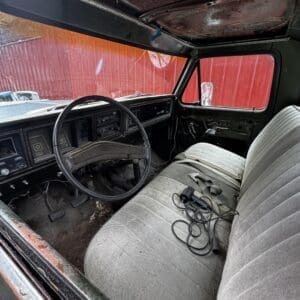 Interior of a rusted, older pickup truck with a gray bench seat, cracked dashboard, and dusty floor; red barn-like exterior visible through the windows.