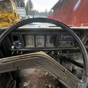 Interior view of a dusty vintage Ford truck cab, steering wheel centered, weathered dashboard with square gauges and switches, dirt floors, and outdoor yard with yellow machinery and a red barn visible through the windshield