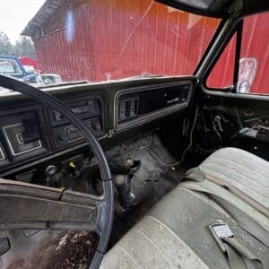 Dusty, worn interior of an old pickup truck with a manual gear shifter, cracked dashboard, and dirt on the floor; a red barn is visible through the windshield outside.