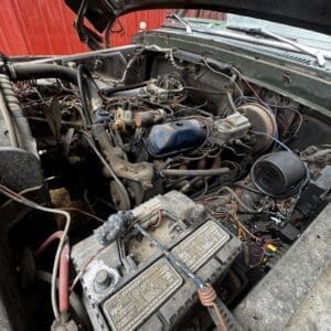 Engine bay of an older vehicle with a dusty battery and exposed wiring under the raised hood in a cluttered, workshop-like area.