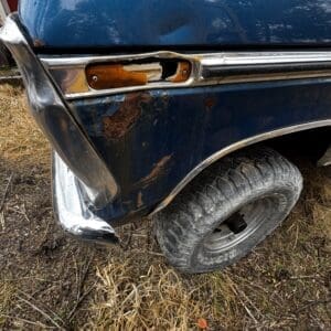 Blue vintage car with rusted body, missing front turn signal, and dented bumper near the wheel on dry grass.