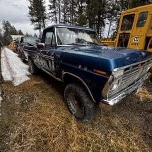 Three-quarter view of a blue vintage pickup truck parked on dry grass with a yellow CAT construction vehicle in the background among pine trees.