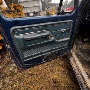 Open blue truck door showing a teal/green interior panel with a manual window crank; muddy ground and yellow construction machinery outside.