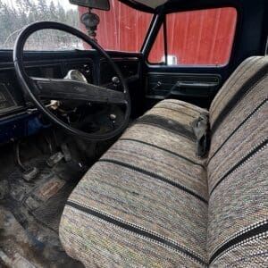 Interior of a dusty vintage truck cabin with a black steering wheel and striped fabric bench seat, outside a red barn and trees.