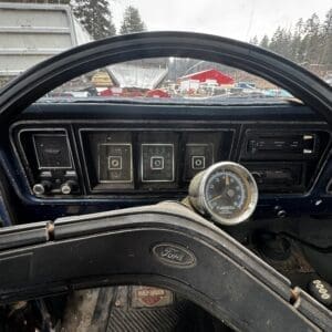 Interior of a vintage Ford truck: three toggle switches on a weathered dashboard, a round gauge near the steering wheel, Ford emblem on the steering column, Harley-Davidson floor mat, snow outside.