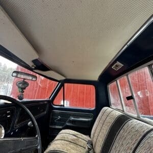 Interior of an old pickup truck with a striped bench seat, black dashboard, and perforated beige headliner; a red structure is seen through the windows.