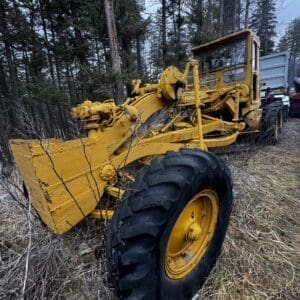 Yellow heavy-duty road grader with a front blade sits on a grassy, forested slope, large tire in the foreground and trees in the background, suggesting a rural maintenance scene.