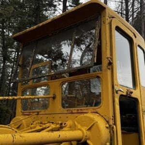 Close-up of a yellow, rusted construction vehicle cab with large windows, standing in a forest.