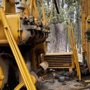 Close-up of a battered yellow construction machine with rusted components and scattered parts in a forested area