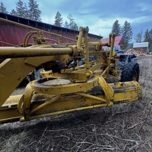 Old yellow farming implement attached to a tractor in a rural yard, rusted and dirty with dry grass around, red barn and wood piles in the background.