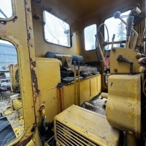 Old, yellow construction vehicle cab interior with rusted panels, worn seat, and dusty controls.