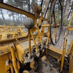 Interior of a weathered yellow heavy machinery cab with multiple levers, pedals, and a large steering wheel; dusty with a forest backdrop beyond the windows.