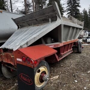 Red Peterbilt dump trailer with a raised metal bed, parked in a junkyard among debris and trees.
