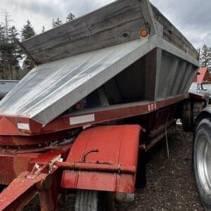 Red dump trailer hitched to a truck with its metal bed raised, showing a wooden side panel and warning reflectors, on a dirt lot with trees in the background.