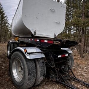 Rear view of a large gray dump trailer with dual rear axles, parked on dirt in a forest.