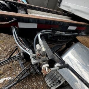 Close-up of a trailer hitch with hydraulic hoses and rusted fittings beneath a flatbed trailer bed