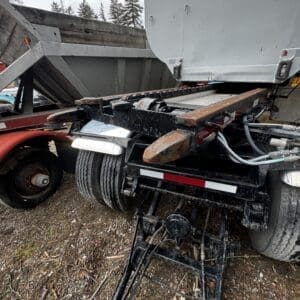 Rear of a heavy trailer with a rusted hitch and tractor-trailer linkage on a gravel lot.