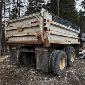 Rear view of a rusty dump truck trailer with dual rear wheels, parked on a rocky, dirt lot among tall trees. Red taillights visible and weathered metal.