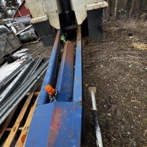 Industrial scrap yard scene with a rusted blue metal beam and a large rusted machine trailer; orange cap and hoses visible, surrounded by metal scraps and wooden pallets.