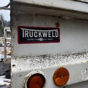 Weathered white truck bed with rust, two orange taillights, and a red-bordered 'TRUCKWELD' decal in a junkyard setting.