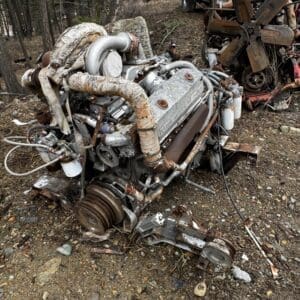 Rusty engine and automotive parts scattered on rocky dirt in a forest clearing, with a white pickup truck in the background.