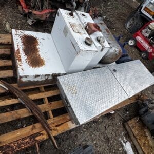 Rusted industrial equipment and a diamond-plate metal box sitting on wooden pallets in a junkyard outdoors.