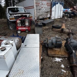 Rusted junkyard scene with an old white pickup, a red portable generator, a diamond-plate metal box, and a weathered trailer among scattered machinery on gravel.