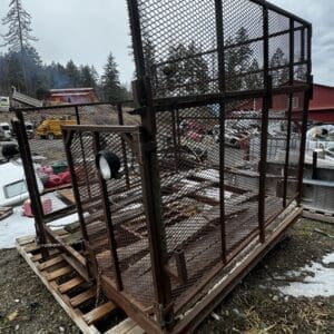 Rusty metal mesh cage on a wooden pallet in a junkyard, with vehicles and debris in the background.
