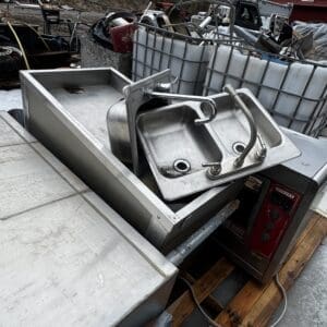 Industrial stainless steel sink and prep station outdoors in a junkyard among salvaged equipment and containers.