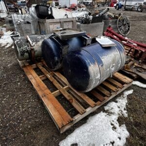 Blue cylindrical tanks on a wooden pallet in a salvage yard, surrounded by scrap metal and old vehicles in the background.