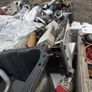 Pile of metal scrap and discarded equipment in a junkyard with trucks in the background on a muddy, snowy lot.