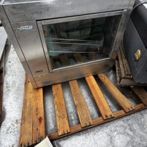 Industrial stainless steel oven with glass door sits on a wooden pallet outdoors in light snow, showing metal racks inside and a Hobart label on the side.
