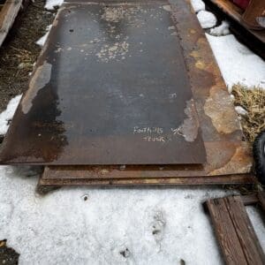 Large rusty metal sheet lying on a rusted frame atop a snowy ground, surrounded by scrap wood and containers.