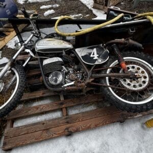 Vintage Yamaha motorcycle with a black number 4 plate, heavily rusted, resting on wooden planks in a snowy, outdoor work area.