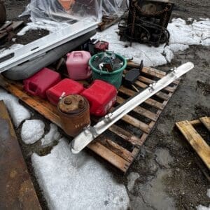 Assorted tools and containers on a wooden pallet in a muddy, snowy yard; includes red plastic gas cans, a green bucket, and a long metal rail.