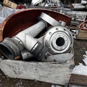Pile of discarded industrial metal parts and pipes in a wooden crate, with a rusted circular duct and metal flange on top in an outdoor salvage yard