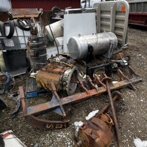 Pile of rusted industrial machinery and tanks in a scrapyard, with a red barn in the background.
