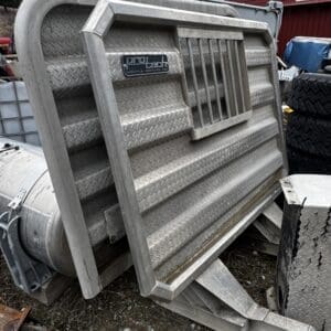 Large diamond-plate metal truck accessory/panel leaning against a frame, with a ProTech badge, outdoors near a red building and tires in the background.