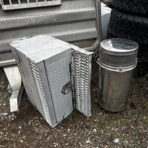 Open diamond-plate metal tool box leaning against a textured metal surface, with a cylindrical metal trash can nearby and stacked tires in the background outdoors.