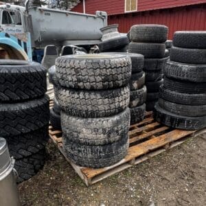 Stacks of used tires piled on a wooden pallet beside a red barn and a blue truck in an outdoor yard.
