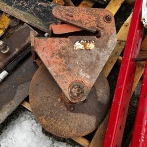 Old rusted industrial pulley block and wheel on a wooden pallet, among scrap metal and a red metal frame.