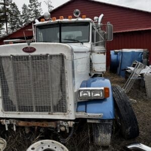 Old white-and-blue Peterbilt truck in a junkyard with rusted grille and wheels scattered around, a red barn in the background.