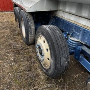 Close-up of a dual-axle trailer with worn tires and blue-painted suspension on dirt ground beside a red building.